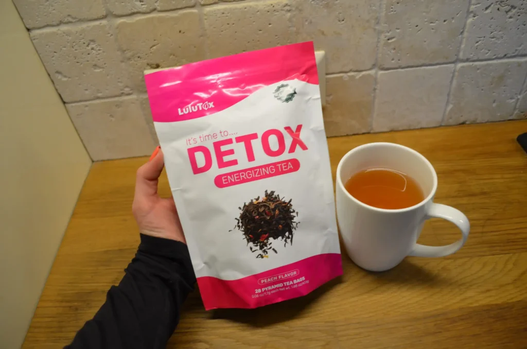 A hand holding a Lulutox Detox Energizing Tea pouch next to a white mug filled with tea.