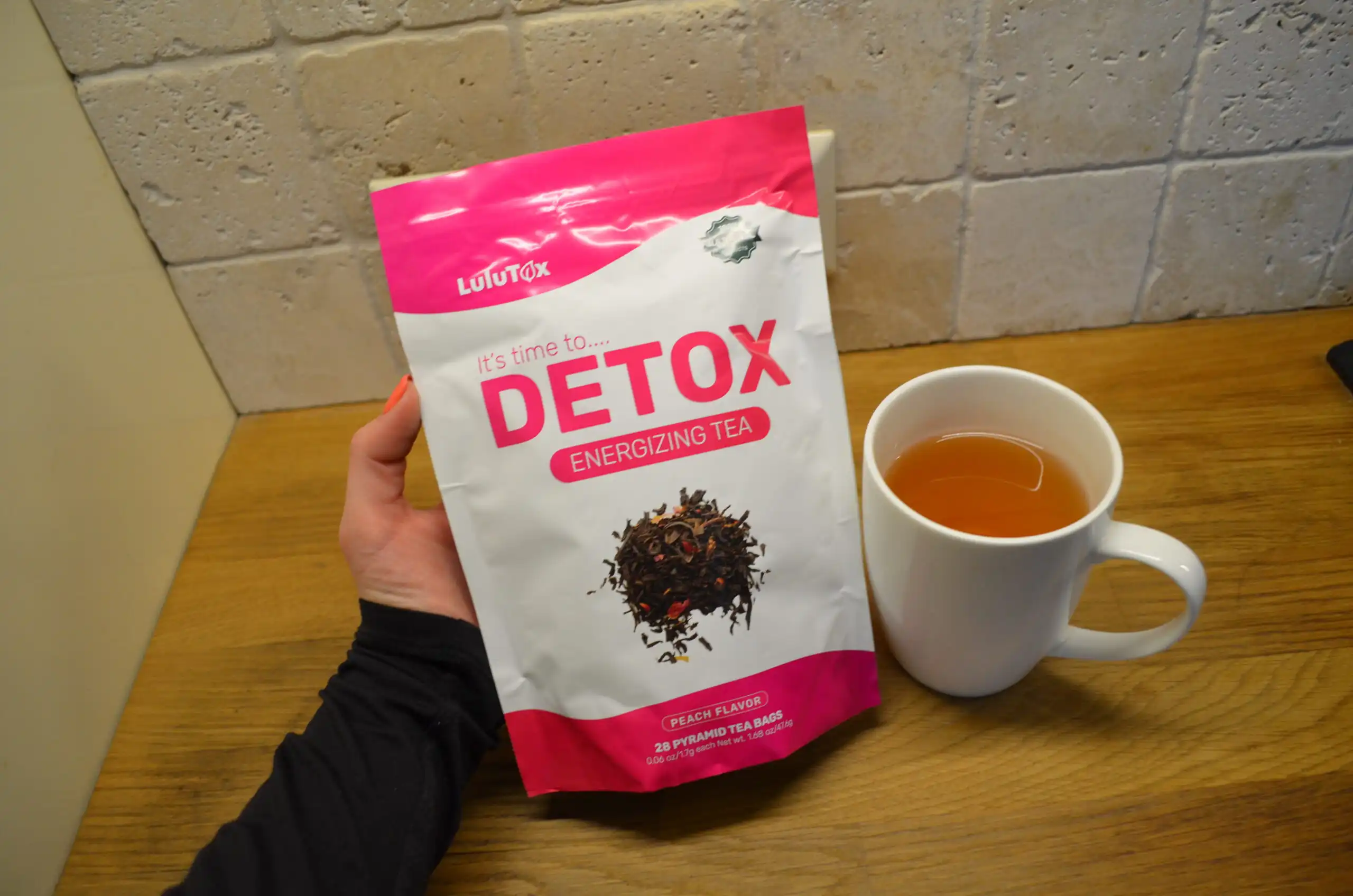 A hand holding a Lulutox Detox Energizing Tea pouch next to a white mug filled with tea.